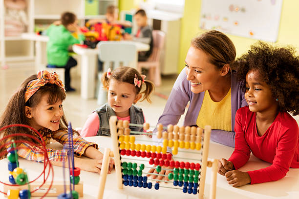 Smiling teacher and little girls talking and playing with educational toys at preschool. Other children are in the background.